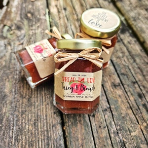 May include: Three small glass jars with gold lids and brown paper labels. The labels have a red apple design and the text "SPREAD THE LOVE", "Turrey & Brenda", "BOURBON APPLE BUTTER", and "APRIL 28, 2024".