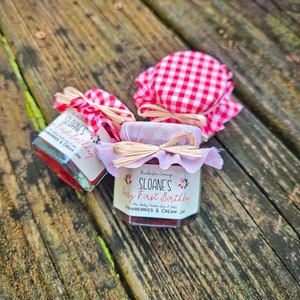 May include: Three small glass jars with red strawberry and cream jam. Each jar is decorated with a pink and white gingham fabric lid, a pink ribbon, and a straw bow. The labels on the jars say "Sloane's First Birthday" and "Strawberries & Cream Jam".