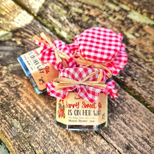May include: Three small glass jars of mixed berry jam with red and white checkered fabric lids tied with a brown ribbon. The labels on the jars say "Berry Sweet Girl is on her way".