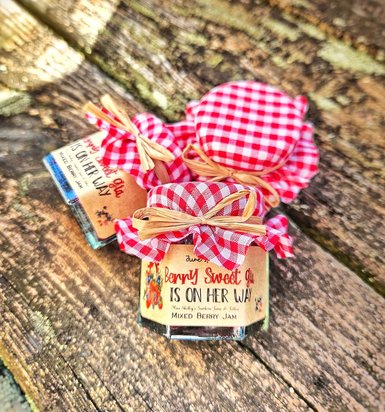 May include: Three small glass jars of mixed berry jam with red and white checkered fabric lids tied with a brown ribbon. The labels on the jars say "Berry Sweet Girl is on her way".