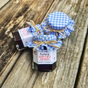May include: Three small glass jars of blueberry lavender jam with blue and white gingham fabric lids and twine tied around the lids. The labels on the jars say "Kiara's Berry First Birthday".