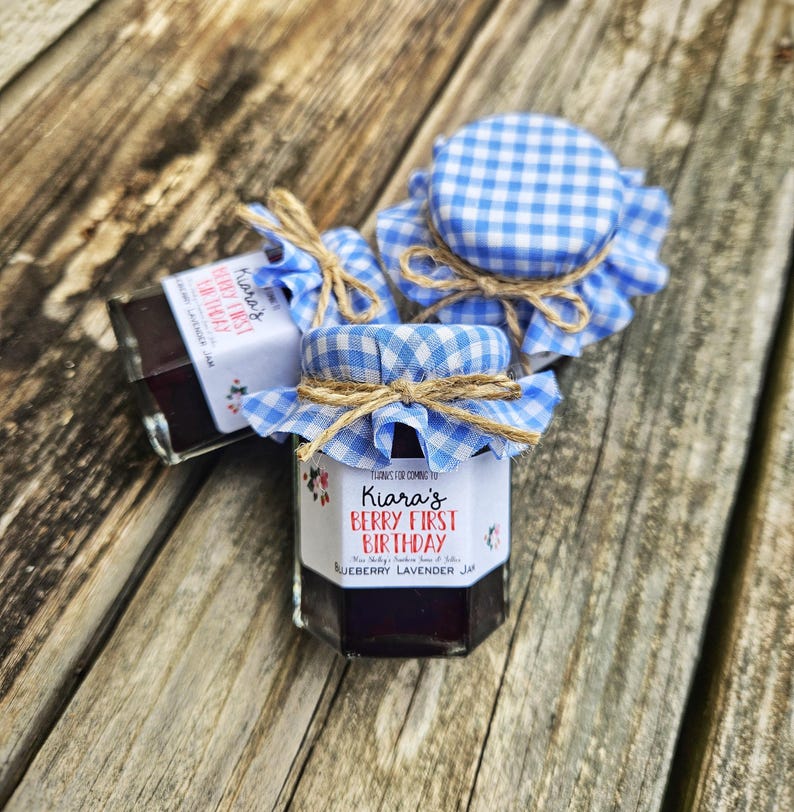 May include: Three small glass jars of blueberry lavender jam with blue and white gingham fabric lids and twine tied around the lids. The labels on the jars say "Kiara's Berry First Birthday".