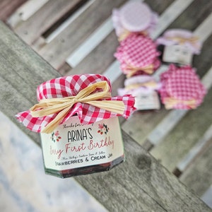 May include: A small glass jar with a red and white checkered fabric lid and a brown ribbon. The jar contains a red and white strawberry jam and has a label that reads "Thanks for celebrating Arina's First Birthday. Miss Shelley's Southern Jams & Jellies. Strawberries & Cream Jam."