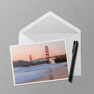 May include: A photograph of the Golden Gate Bridge at sunset is displayed on a white card, placed inside an open white envelope. A black pen rests beside the card and envelope on a gray surface. The bridge is red, and the water is blue.