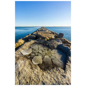 May include: A stone pier extends into a calm, blue ocean under a clear, bright sky. The pier is constructed of large, weathered rocks. A small pool of ice sits on the pier's surface, reflecting the sky. The scene evokes a sense of tranquility.