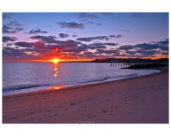 Puede incluir: Un atardecer en la playa con un sol naranja brillante que se refleja en el agua. El cielo cambia de naranja a morado, con nubes oscuras. La playa de arena está en primer plano, con un muelle y edificios a lo lejos.