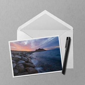 May include: A photograph of a coastal landscape with a vibrant sunset over the ocean. The image is on a white card, placed in an open white envelope, with a black pen resting beside it. The scene includes rocks and water.