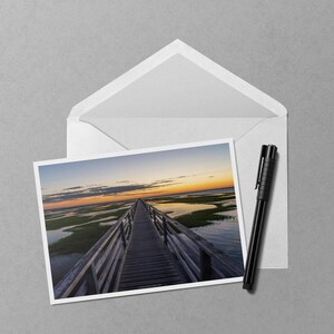 May include: A photograph of a wooden boardwalk extending over water and marshland towards the horizon at sunset. The image is on a white card, inside a white envelope, with a black pen to the side.