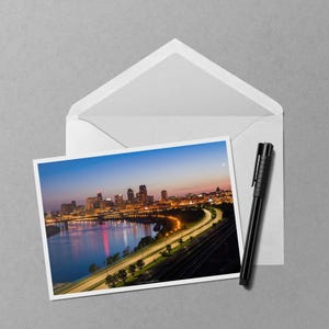 May include: A photograph of a cityscape at dusk, featuring a river, illuminated buildings, and a highway with car lights. The photo is inside a white envelope, with a black pen resting beside it.