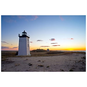 May include: A white lighthouse with a black top stands on a sandy beach under a colorful sunset. A small building and a hill are in the background. The sky is a mix of blue, pink, and orange.