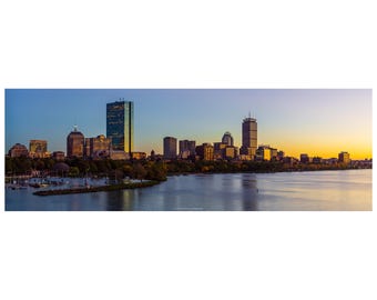 May include: Panoramic view of a city skyline at dusk. The image features a body of water in the foreground, with a cityscape of buildings and skyscrapers in the background. The sky transitions from blue to orange and yellow.