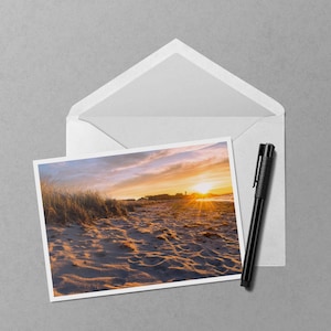 May include: A photograph of a beach scene at sunset, featuring a vibrant orange and yellow sky. The image is in a white frame, placed in a white envelope, with a black pen resting beside it. The scene includes sand, grass, and a distant shoreline.