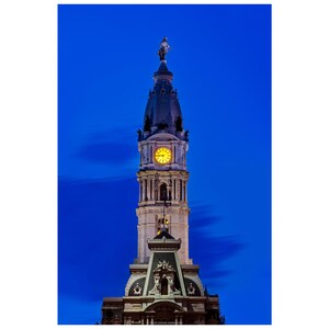 May include: A tall, ornate building tower against a deep blue sky. The tower features a clock with a yellow face, architectural details, and a statue at the top. The image is well-lit, highlighting the building's details.
