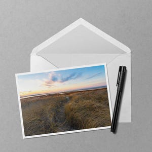 May include: A landscape photograph of a coastal scene with tall, dry grasses and a path leading to the horizon. The sky is a mix of blue, orange, and pink. A white envelope and a black pen are next to the photo.