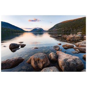 May include: A scenic landscape photograph of a lake surrounded by mountains. Large, rounded rocks are in the foreground, with clear water revealing smaller rocks beneath. The sky has soft pink and orange hues.