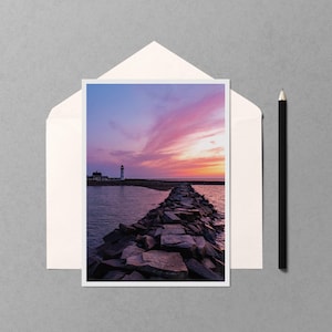 May include: A photograph of a coastal scene featuring a stone pier leading to a lighthouse under a vibrant sunset sky. The image is in a white frame, placed inside an envelope, with a black pencil to the side.