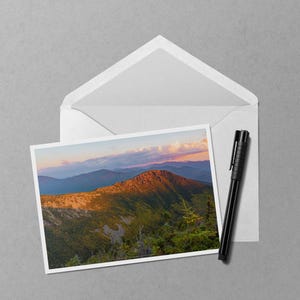 May include: A landscape photograph of a mountain range at sunset, displayed in a white envelope. A black pen rests beside the envelope. The photograph shows a mountain with golden sunlight and a blue sky.