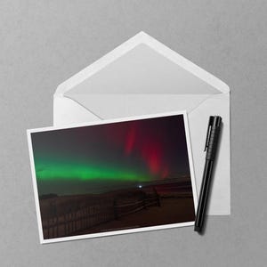 May include: A photograph of the aurora borealis, with vibrant green and red hues illuminating the night sky. The image is on a white card, placed inside an open white envelope, with a black pen resting beside it. The scene evokes a sense of wonder.