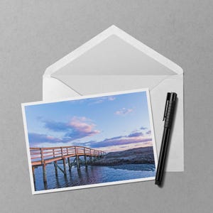 May include: A white envelope with a landscape photograph and a black pen. The photograph shows a wooden bridge over water under a blue and pink sky. The pen is on the right side of the envelope.