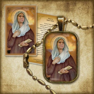 May include: A gold pendant necklace with a glass charm featuring a painting of Saint Adele. The charm has a brown background with a golden halo around the saint's head. The saint is wearing a brown robe and a white veil. She is holding a book in her left hand and her right hand is on her chest. The pendant is hanging from a gold chain with oval-shaped links.