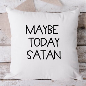 Pu&ograve; includere: Cuscino quadrato bianco con la scritta "MAYBE TODAY SATAN" in nero, carattere sans-serif. Il cuscino &egrave; su una superficie di legno chiaro con uno striscione di tessuto beige sullo sfondo. Un cuscino decorativo.