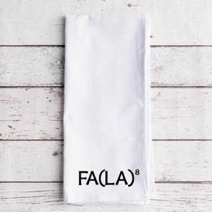 May include: White folded kitchen towel with the text "FA(LA)⁸" printed in black. The towel is made of a soft, absorbent material and is perfect for drying dishes or wiping surfaces. The towel is displayed on a white wooden surface.