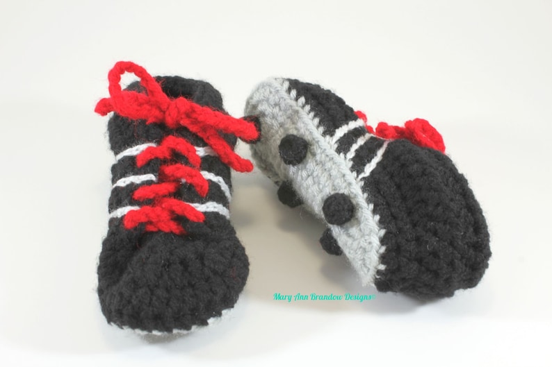 May include: A pair of crocheted baby booties in the shape of soccer cleats. The booties are black with white stripes and red laces. The soles are gray with black dots.