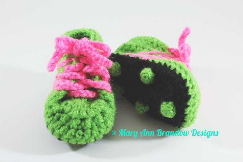 May include: A pair of crocheted baby shoes in green and pink. The shoes have a black sole with raised dots. The shoes are tied with pink laces.