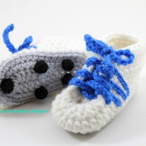 May include: A pair of crocheted baby shoes in white and gray with blue laces. The shoes have a simple design and are perfect for a baby's first steps.