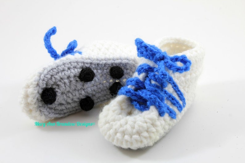 May include: A pair of crocheted baby shoes in white and gray with blue laces. The shoes have a simple design and are perfect for a baby's first steps.