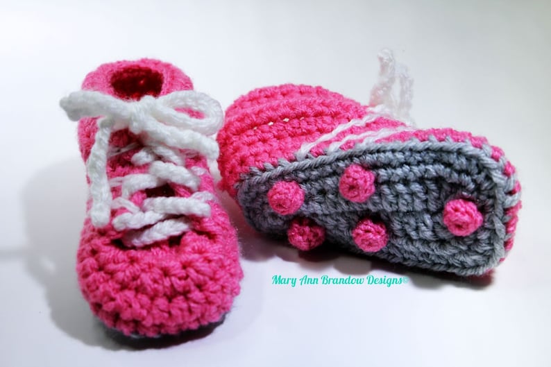 May include: A pair of pink and gray crocheted baby booties with white laces. The booties have a textured sole with pink dots.