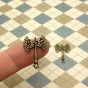 以下が含まれることがあります： 詳細なデザインが施されたアンティークブロンズの斧チャーム2個。斧の頭は湾曲しており、表面はテクスチャ加工されています。チャームの上部にはループがあり、ネックレスやブレスレットに取り付けることができます。