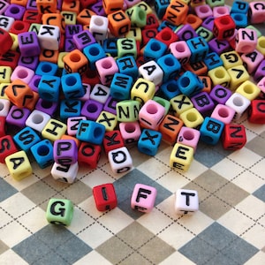 Può includere: Un mucchio di perline di plastica colorate a forma di lettere, con le lettere G, I, F e T scritte su una superficie a scacchi.