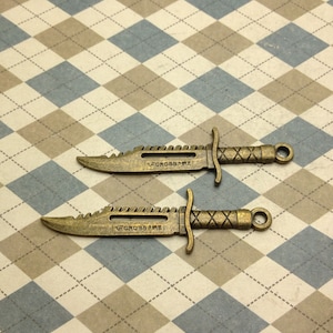 May include: Two bronze-colored miniature knives with serrated blades and cross-shaped handles. The text "V CROSS FIRE" is etched on the blade. The knives have a loop at the end of the handle.
