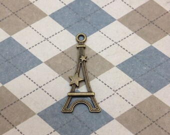 Puede incluir: Un amuleto de metal de color bronce con forma de Torre Eiffel con tres estrellas en la parte superior. El amuleto está sobre un fondo a cuadros blanco y azul.