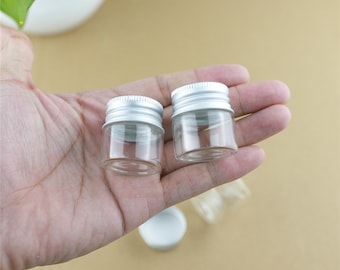 May include: Two small, clear glass jars with silver metal screw-on lids are held in a person's hand. The jars are empty and appear to be new. A white lid is visible in the background.