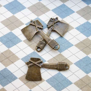 以下が含まれることがあります： ロープのディテールが付いたアンティークブロンズの斧チャーム3つ。チャームは白と青の市松模様の背景に十字形に配置されています。