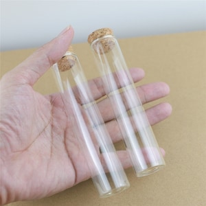 Può includere: Due provette di vetro trasparente con tappi di sughero. Le provette sono alte circa 15 cm e hanno un diametro di 2,5 cm.