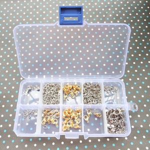 Puede incluir: Una caja de almacenamiento de plástico transparente con tapa azul, que contiene ocho compartimentos llenos de diversos hallazgos de joyería de metal plateado y dorado.