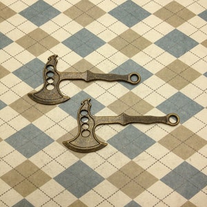 以下が含まれることがあります： 装飾的なデザインが施されたアンティークゴールドの金属製の斧チャーム2個。チャームの上部には、ネックレスやブレスレットに取り付けるための穴があります。