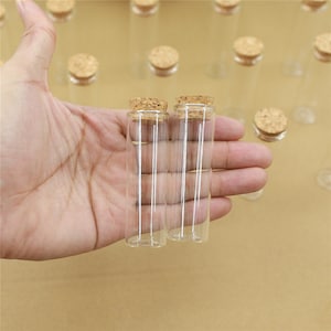 Könnte beinhalten: Zwei klare Glasfläschchen mit Korkstopfen. Die Fläschchen werden in einer Hand gehalten.