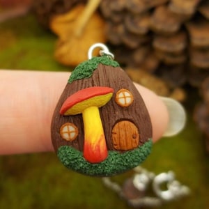 May include: A small, brown, mushroom-shaped house pendant with a red and yellow mushroom growing out of the roof. The house has two windows and a small door. The pendant is made of polymer clay and is attached to a silver chain.