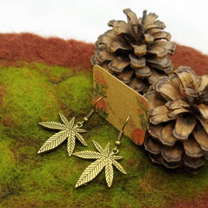 May include: A pair of gold-toned earrings featuring a marijuana leaf design. The earrings are displayed on a brown card with a floral pattern and a pine cone.