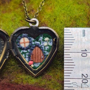 May include: Heart-shaped locket necklace with a miniature fairy house scene inside. The bronze-toned locket opens to reveal a tiny door, window, and mushrooms set against a stone wall and green moss. The locket is suspended from a matching chain.