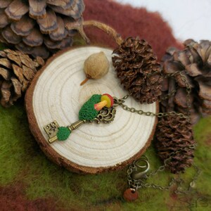 May include: A green and brown key necklace with a small red and yellow mushroom charm. The necklace is on a wooden surface with pine cones.