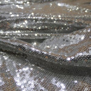 May include: Close-up of a silver sequin fabric. The fabric is draped, creating folds and shadows that highlight the texture and sparkle of the sequins. The sequins are round and closely spaced.