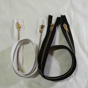 Puede incluir: Primer plano de varias cremalleras en blanco y negro con dientes y tiradores de metal dorado. Una cremallera blanca está enrollada. Las cremalleras están sobre un fondo blanco. Para proyectos de costura.