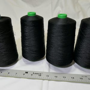 May include: Four black spools of thread with green tops are arranged in a row. A metal ruler lies in front of the spools. The spools are cylindrical and filled with tightly wound thread, suitable for sewing and crafting projects.