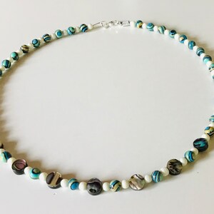 May include: A necklace with a mix of white, blue, and iridescent beads. The necklace has a silver clasp and is composed of small white beads interspersed with larger, patterned beads in shades of blue, green, and iridescent colours.