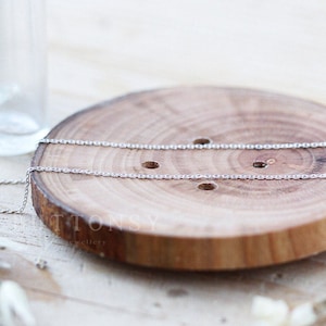 May include: Two delicate silver chain necklaces are displayed on a round, wooden coaster. The chains are thin and have a simple, elegant design. The coaster is made of light brown wood with a natural grain.